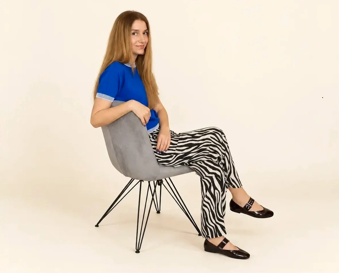 Aleksandra smiling, sitting sideways on a grey chair with wire legs in a blue top and zebra-print trousers