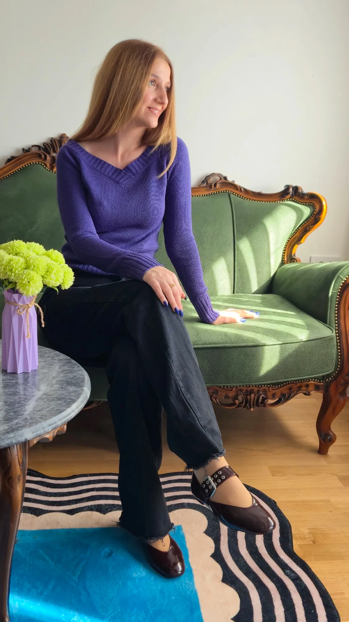 Aleksandra sitting on a green velvet sofa in a purple top, beside a vase of green flowers and a striped rug